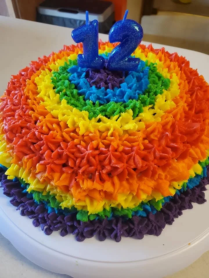 Rainbow-frosted round birthday cake topped with a blue number two